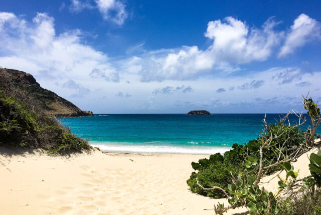 Plage tropicale de sable blanc et d'eau turquoise, entourée de collines verdoyantes.