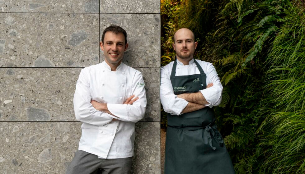 Two chefs in white coats, standing with arms crossed, one by a concrete wall, the other by greenery.